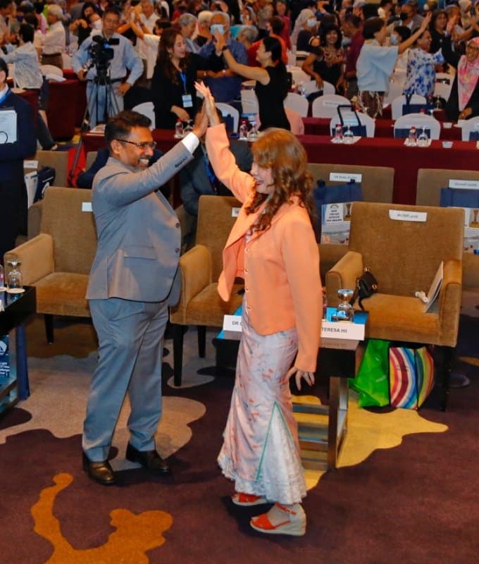 Teresa Hii in a peach jacket high-fives a colleague while conference participants stand and clap behind them